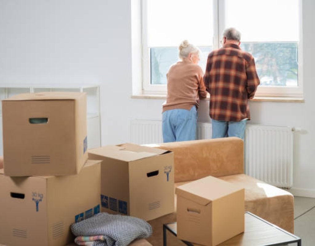 Heap of cardboard boxes with senior couple looking through window in the background. Senior couple moving house.