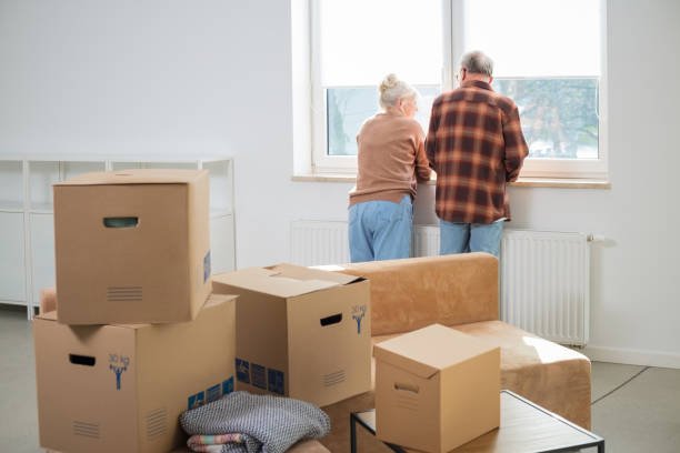 Heap of cardboard boxes with senior couple looking through window in the background. Senior couple moving house.